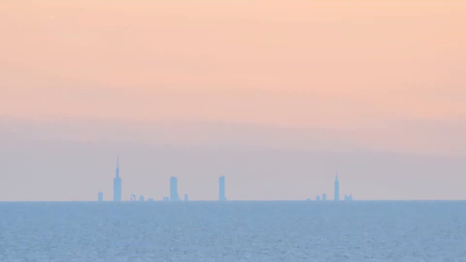 A frame of the time-lapse: a pale evening sky over Lake Michigan, and along the water horizon a thin row of four tower silhouettes, the Willis with its masts at left and the Hancock at right, standing directly on a level line
