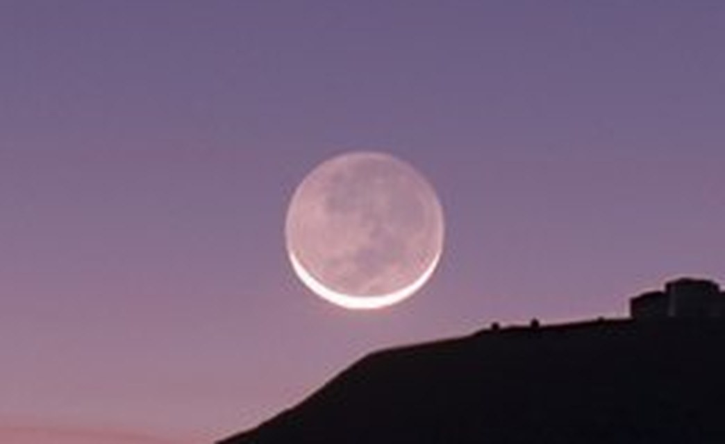 Detail crop of the same Moon: a brilliant thin crescent along the lower edge, and the entire remaining disc clearly visible as a soft grey circle with dark maria discernible on it, lit by earthshine.