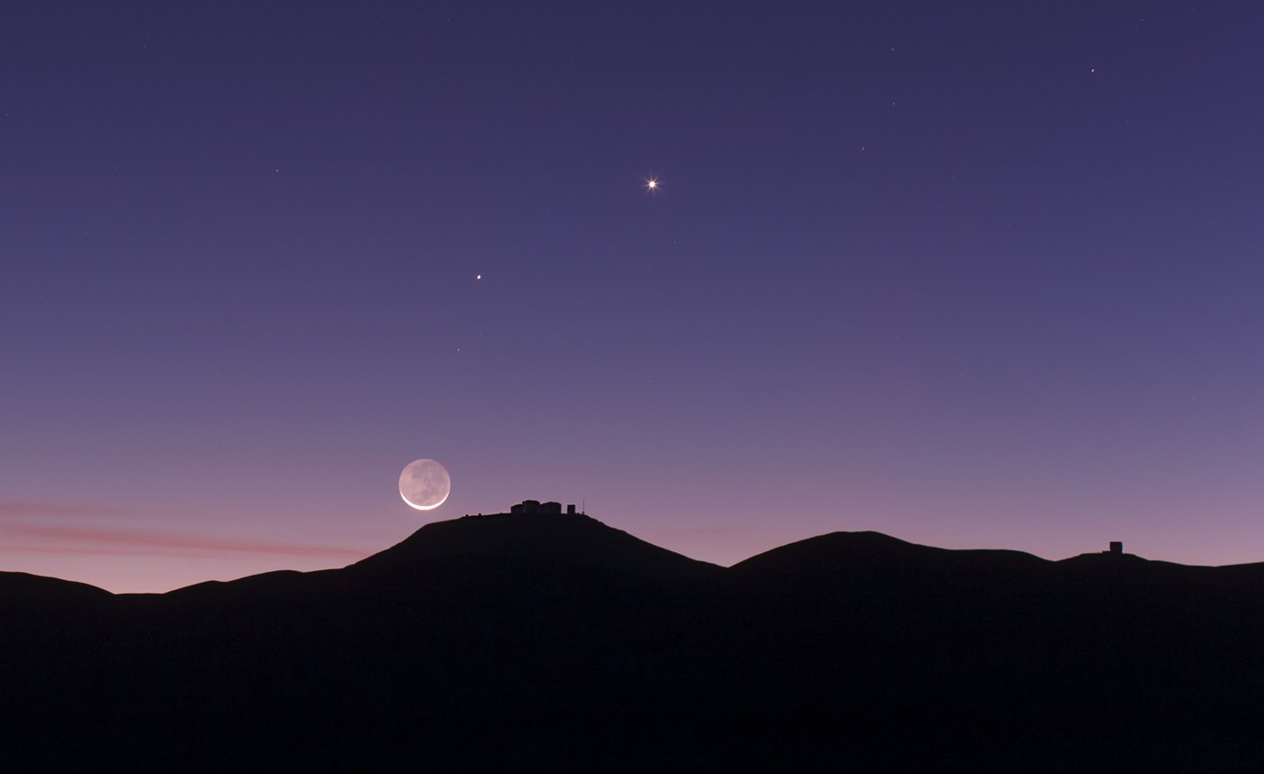 A thin crescent Moon low over the darkened ridge of ESO's Paranal Observatory at twilight, with Venus and Mercury as bright points in a deep violet sky.