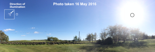 A wide panorama of a Scottish field on a clear afternoon, captioned Photo taken 16 May 2016. Near the left edge a small square box marks the Moon, and a magnified inset above it, labelled Direction of illumination, shows a gibbous Moon with a white arrow drawn out of its lit side pointing up and to the right. Far over at the right edge a black circle marks the Sun inside its own glare, noticeably lower in the frame than the arrow points.