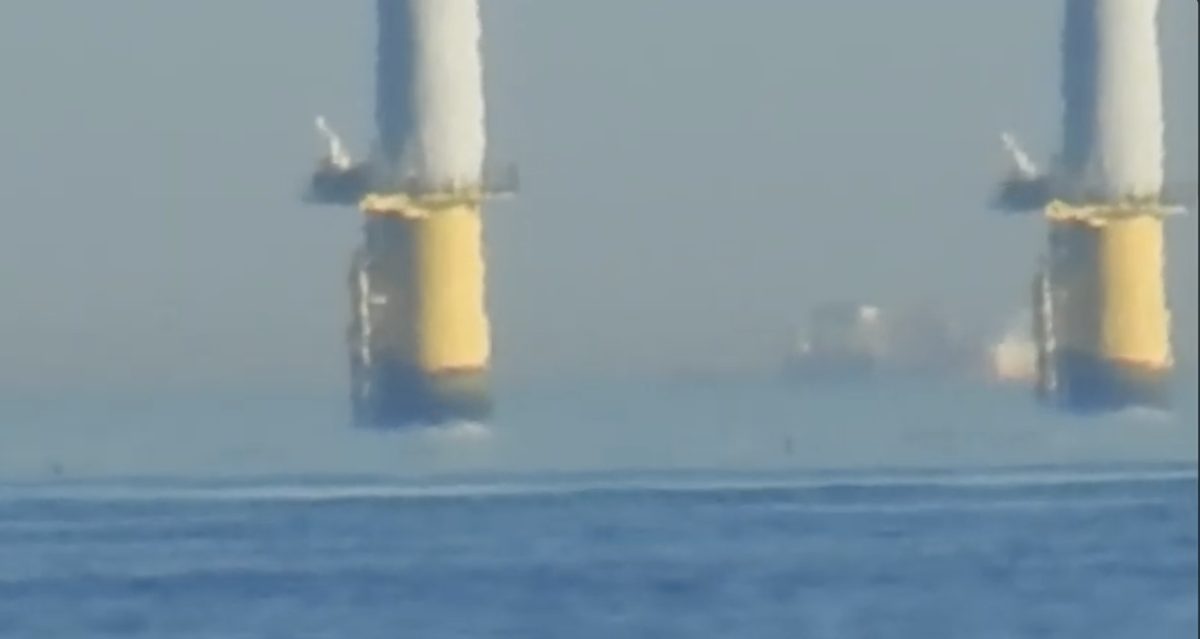 Close view of two turbine monopiles with yellow bases; the bases dissolve into a bright hazy band, distorted and smeared, with a faint ship floating in the haze between them.