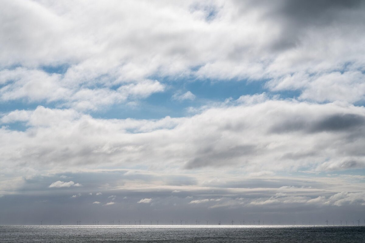 Rampion wind turbines seen as a thin, low silhouette strung along the horizon from the Sussex coast, under an ordinary overcast sky with no visible haze band or lofting.
