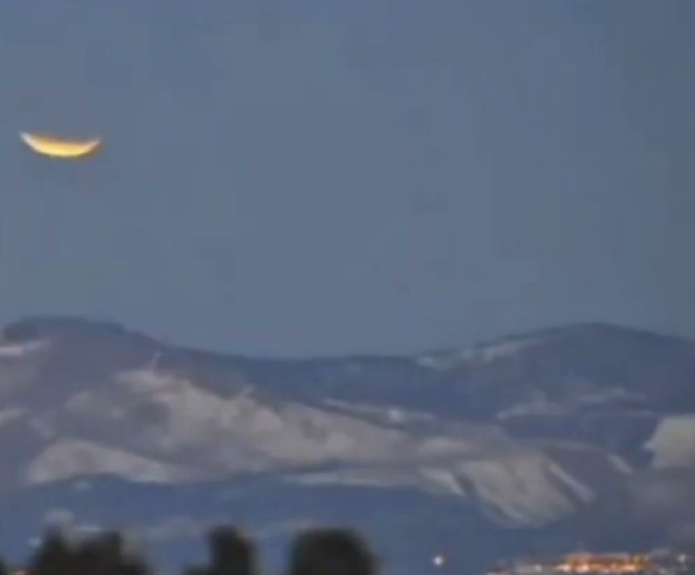 A still from the Santa Fe segment of the video: a thin bright crescent, lit along its lower edge and dark across the top, hanging in a grey-blue twilight sky above a long snow-streaked mountain ridge, with the lights of a town on the plain at lower right.