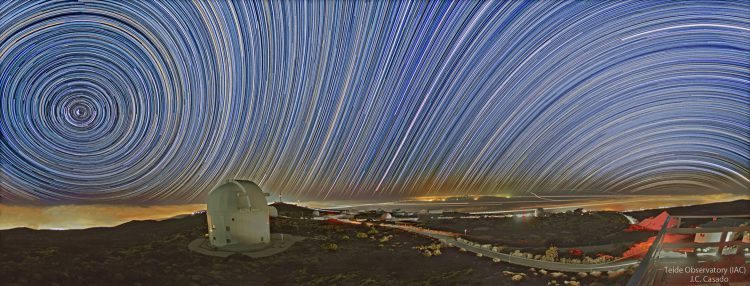 Long-exposure star-trail panorama over Teide Observatory, Tenerife: a tight anticlockwise spiral around the north celestial pole on the left, trails straightening toward the east in the middle, and curving the other way toward the right.